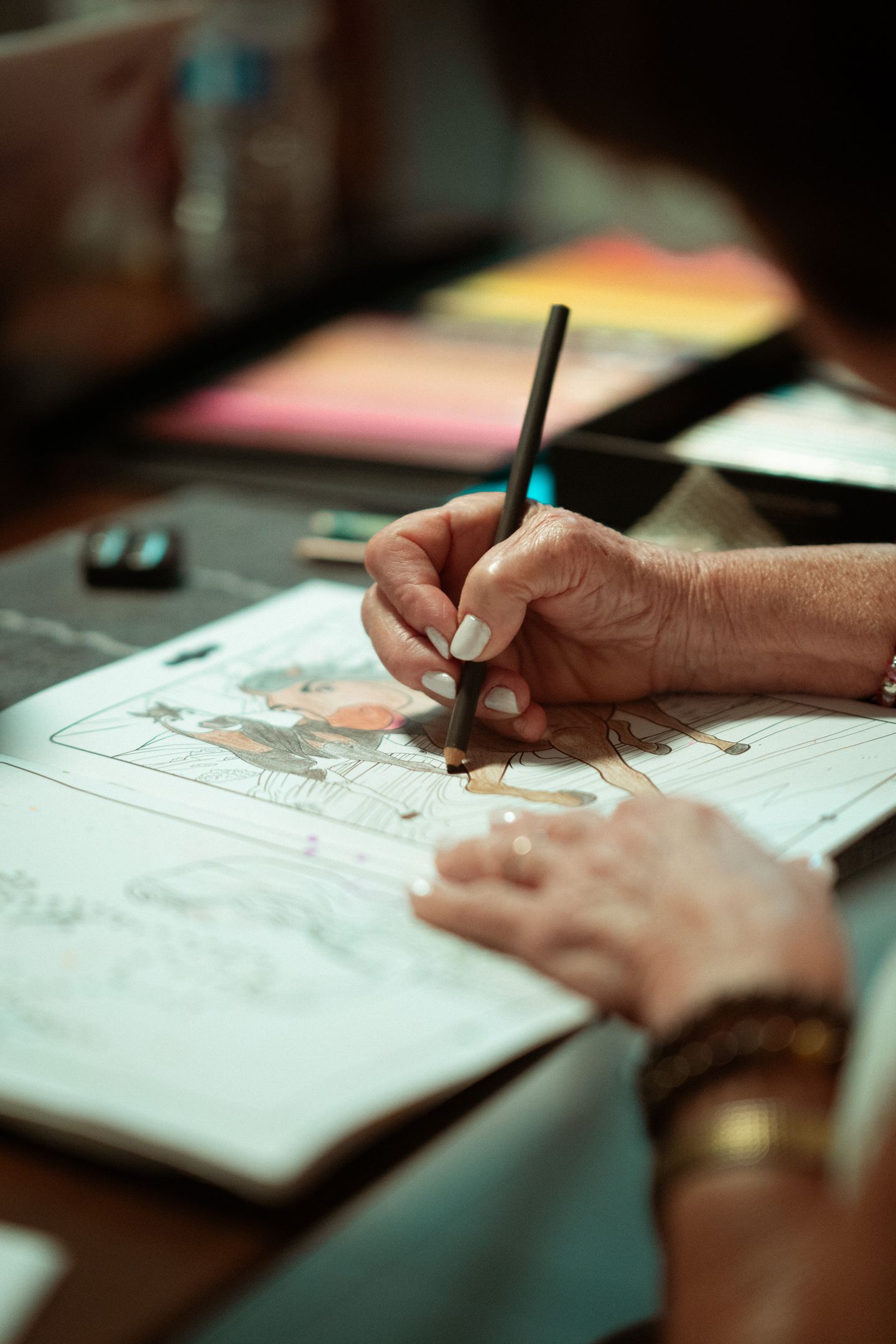 An adult colors an illustrated page with a pencil at a table.