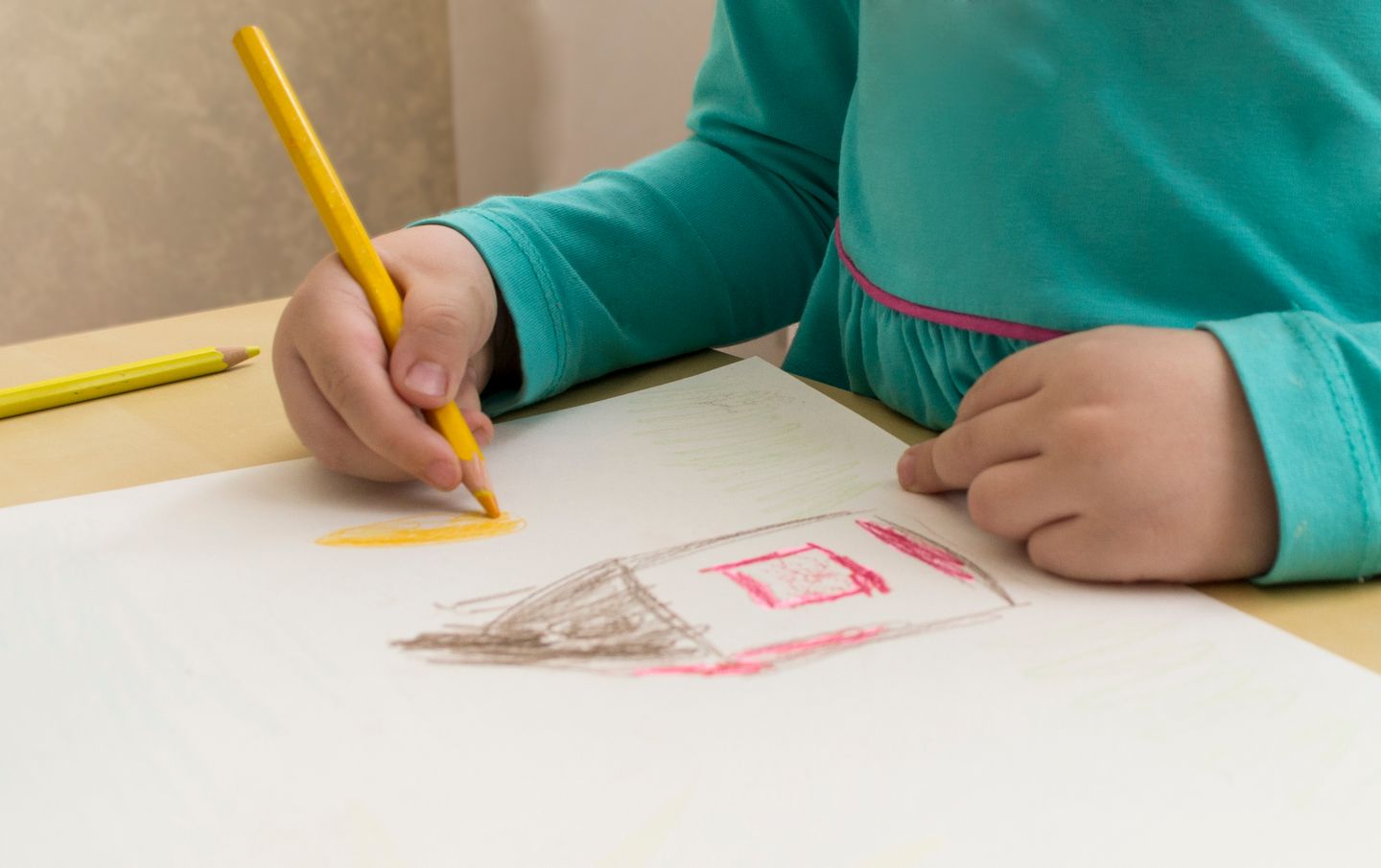 A child in a turquoise top draws a house with a yellow pencil.