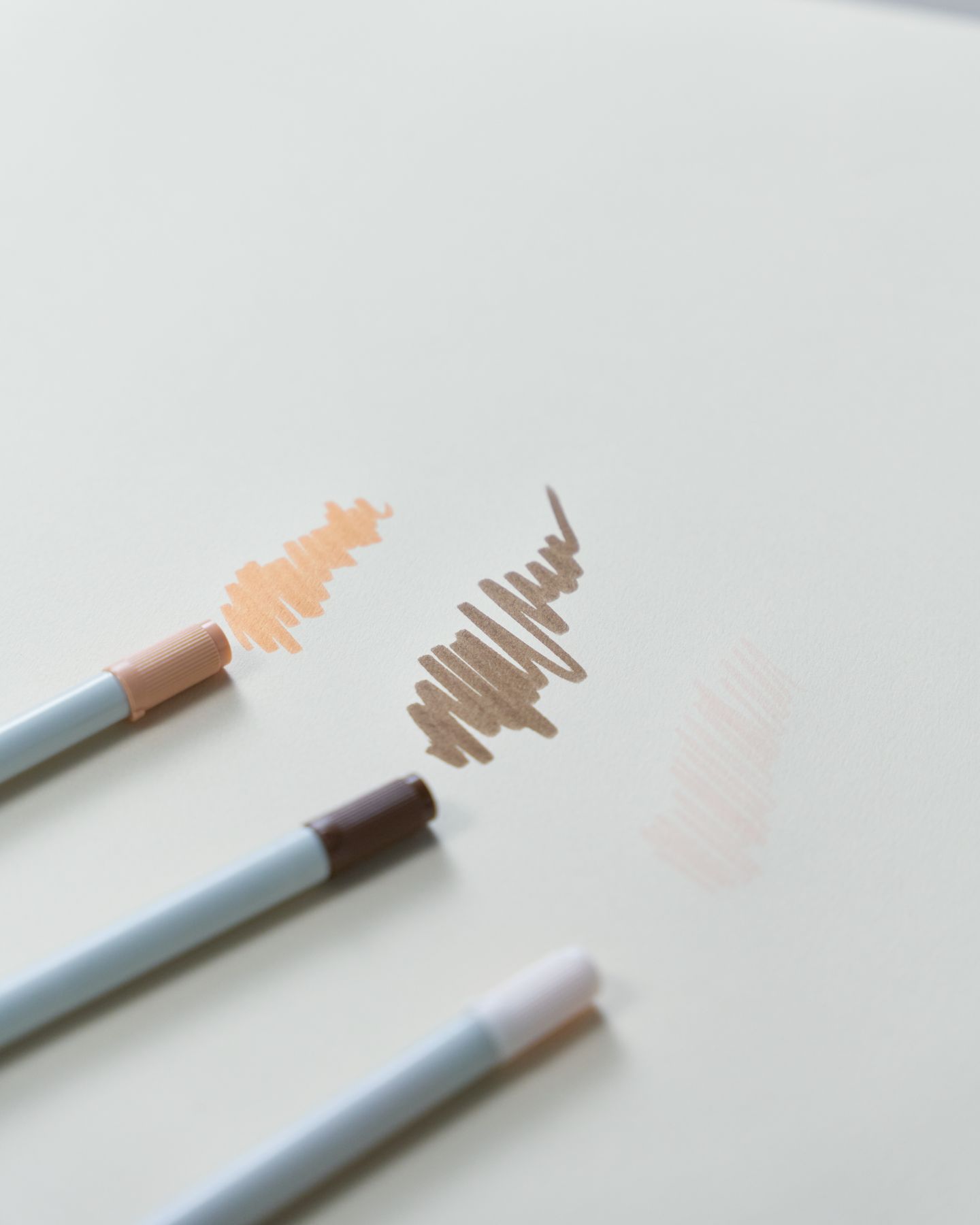 Three coloring pens beside peach, brown, and pink marks on white paper.